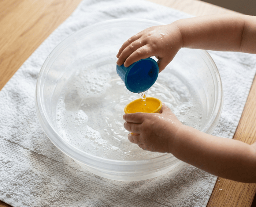 Shallow dish tub of soapy water with plastic cups on a towel for sensory play